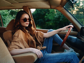 Young woman in sunglasses sitting in a vintage car, wearing a casual outfit of a brown shirt and...