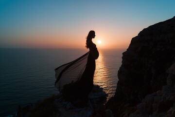 Silhouette of pregnant woman in flowy gown, standing on cliff at sunset, dramatic ocean view and colorful sky, romantic moody maternity portrait with movement and elegance

