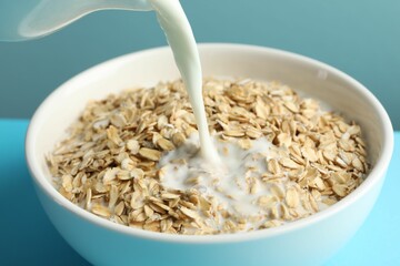 Pouring milk into bowl with oatmeal at light blue table, closeup