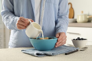 Woman pouring milk into bowl with oatmeal and blueberries at beige textured table, closeup