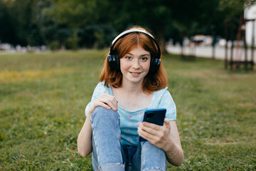 Redhaired teenage girl with freckles in wireless headphones and smartphone sitting on the grass