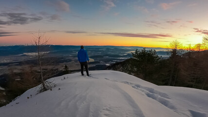Winter hiker in blue jacket standing on snow-covered peak of Mallestiger Mittagskogel, Austria, watching dramatic sunset over Rosental valley and remote Karawanks mountain range with golden fierce sky