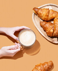 Hand holding cup with cappuccino coffee on orange background with fresh croissants on wooden plate.