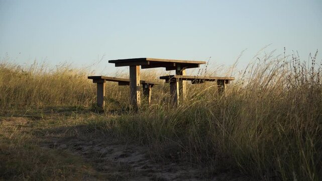Footage of a wooden table and two banches standing on a hill,  grass waving on a wind. Sunset  time, late summer,  calm and mild mood,