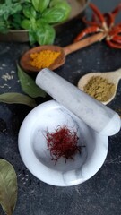 A small amount of real red saffron in light-gray marble mortar. In the background - spices, bay leaves, chili pepper and basil.