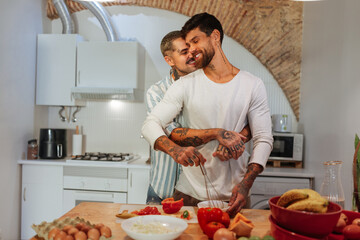 Smiling, tattooed men embrace while cooking healthy food in a stylish kitchen,