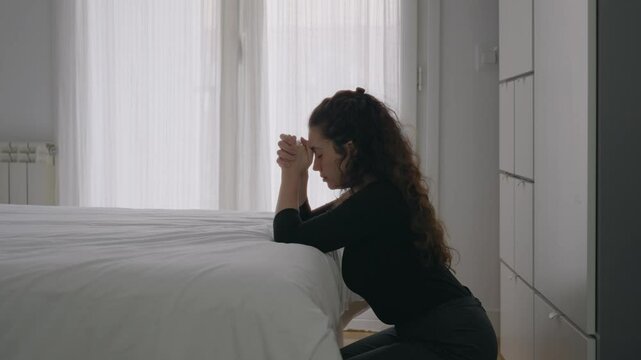Female kneeling by the bedside and praying with eyes closed and hands clasped. Religious woman inside apartment during day time in prayer or worship. Holding hands together speaking to herself