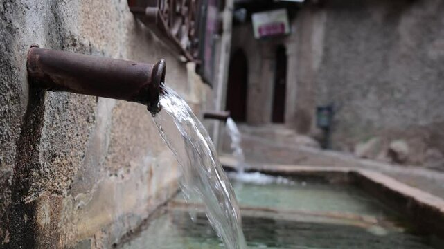 Depicts a rustic water spout from which clear water flows, surrounded by cobblestone pathways and textured walls in a charming village setting.
