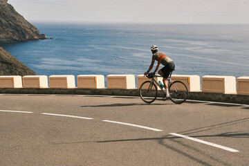 Fototapeta premium Cyclist rides along scenic coastal road. Stunning ocean views and bright sunlight create a picturesque backdrop for this active lifestyle image. Perfect for travel, sports, and healthy living themes.