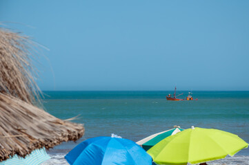 An old fishing boat sailing in front of a beach in Mar del Plata covered with colorful umbrellas
