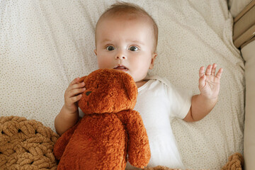 Adorable baby lying in crib with cute little toy teddy under knitted blanket. Time for sleep. Sweet...