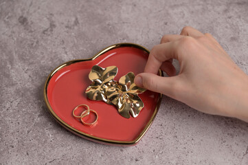 Couple of wedding golden rings and earrings on red heart shape ceramic plate with a gold rim. Woman's hand touch to jewelry. Gray concrete background. Valentine's Day, marriage, wedding