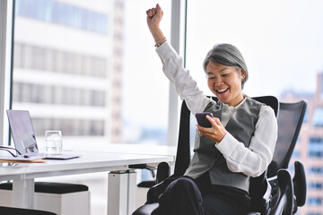Excited mature older Asian business woman winner executive looking at cell phone celebrating...