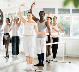 Group of multinational men and women practicing third ballet position at barre in dance studio