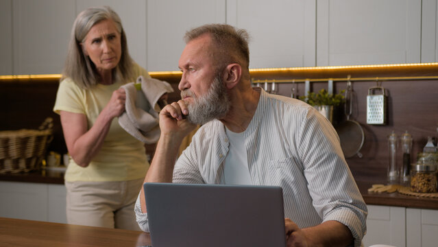 Retired married family frustrated old senior man pay bills online remote work at kitchen table worried about loan debt pension annoyed Caucasian woman wife washing dishes scold husband home quarrel