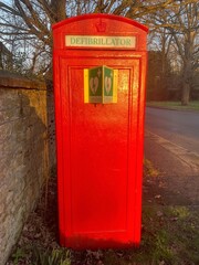 A repurposed classic British red telephone box, now housing a defibrillator, set against a rural stone wall and tree-lined road in warm evening light.