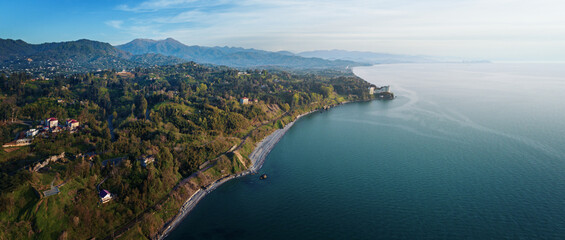 A wide aerial panorama showcasing a coastal Adjara region in Georgia with lush green hills, a winding shoreline, and a calm blue sea, with a luxurious hotel on a cliff and distant mountains