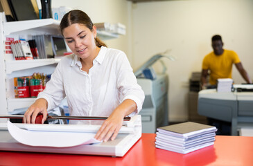 Fototapeta premium Cheerful young woman in white shirt smiling at the camera and using paper cutter in the printing house