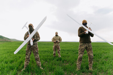Three military personnel stand in a lush field, each holding a drone. They exhibit readiness for an...