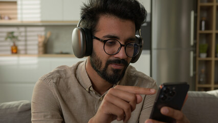 Happy Arabian Indian man in headphones listen music at home kitchen male millennial rest on couch...