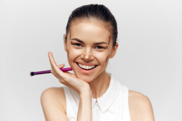 Smiling young woman with makeup brush showcasing beauty and confidence in a light, minimalistic...