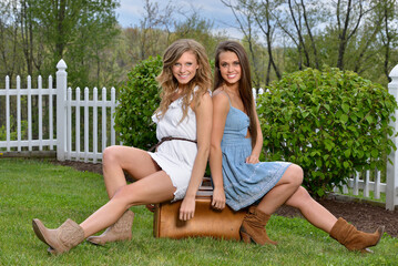Two beautiful women pose together in dresses with vintage suitcase - sitting on suitcase with boots