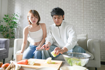 A Japanese pregnant woman in her 20s, wearing a dress that shows her stomach, sits on a sofa in her apartment and her husband cooks a hot pot dish.