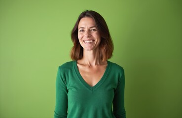 Fototapeta premium Middle-aged woman smiles warmly in studio portrait. She wears green sweater against green background. Happy expression. Casual attire. Possible model or pro. Mid-life beauty. Attractive. Studio shot.