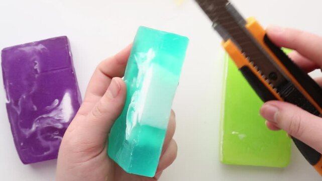 Bright turquoise soap being cut with precision, resulting in clean rectangular pieces. The background features yellow and green soap blocks. Part 1 of 2