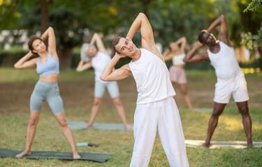 Athletic young man in comfortable sportswear performing Tiryaka Tadasana, or Bending Tree Pose, during group yoga session in green city park on summer day