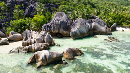 Large granite boulders sit along the turquoise waters with lush greenery and clear skies. Seychelles, La Digue. Anse Source d'Argent.