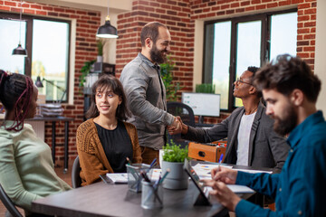 Caucasian man greets african american manager during meeting in brick wall workspace. Businessmen shaking each others hands at table as their coworkers prepare for group discussion in startup office.