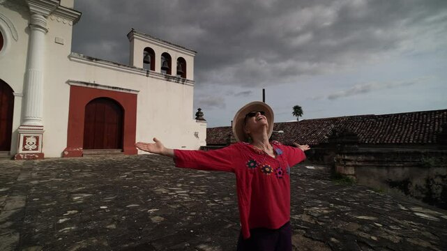 A woman stands with arms outstretched in front of a historic convent in Granada, Nicaragua, embracing the beauty of her surroundings and the cloudy sky.