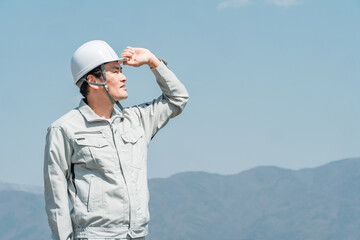 青空の下に立つヘルメットを被った作業着姿の男性（建築・土木・建設）
