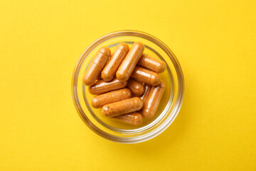 Turmeric capsules in glass bowl on yellow background, top view