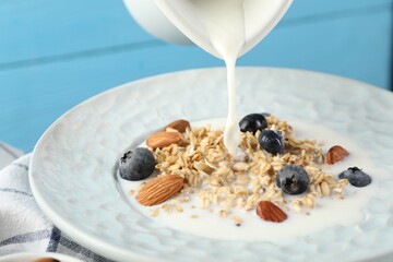 Pouring milk into bowl with oatmeal, blueberries and almonds at light blue table, closeup