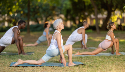 Fit barefoot senior woman in comfortable sportswear practicing yoga at group session on green glade in summer park, performing Anjaneyasana, or low lunge Crescent Moon Pose