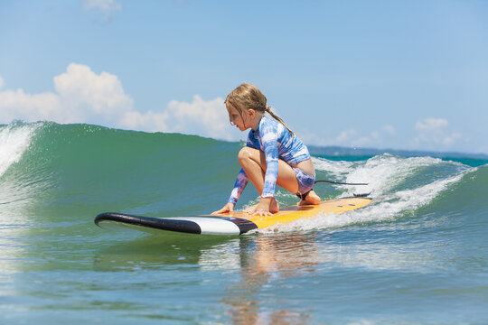 Happy baby girl - young surfer ride on surfboard with fun on sea waves. Active family lifestyle, kids outdoor water sport lessons and swimming activity in surf camp. Summer vacation with child.