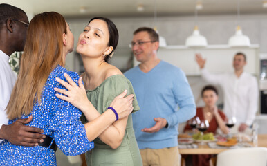 Hospitable Asian woman warmly greeting and kissing female friend arriving for home get-together with food and drinks on table in background..