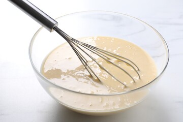 Whisk and bowl of dough on white marble table, closeup