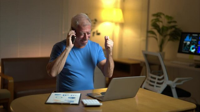 A man is talking on his cell phone while sitting at a desk with a laptop and a stack of papers
