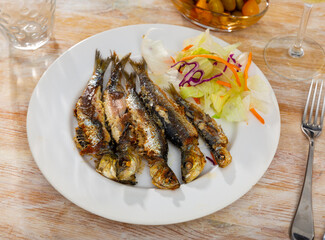 Just cooked fried sardines served on plate on wooden table with lettuce and carrot salad.