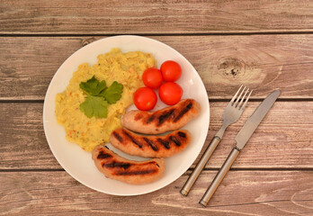 Grilled sausages with mashed potatoes and cherry tomatoes on a wooden table, cutlery nearby.