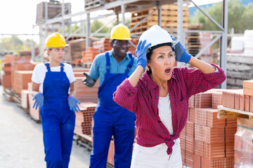 Three builders standing in outdoor construction material storage. Taskmaster in white hardhat amazed by something she just saw.