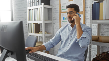 Young man with beard and glasses working in modern office on computer and phone showcasing a...