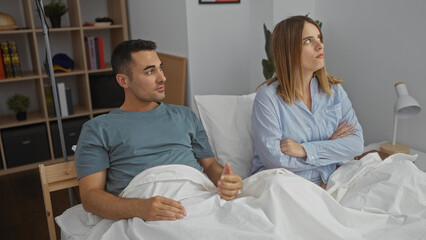 Couple sitting in bed at home with man looking concerned and woman appearing upset in a cozy bedroom setting depicting relationship tension indoors.