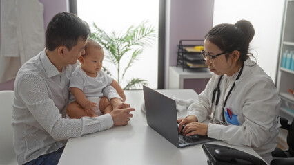 Father with baby boy visiting female pediatrician in hospital clinic room, discussing health with laptop on desk.