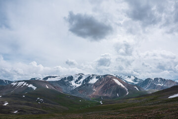 Dramatic alpine view from stony grassy hill to large snow-capped mountain range under cloudy gray sky. Beautiful mountain landscape with green rock hills against high snowy mountains in rainy weather.