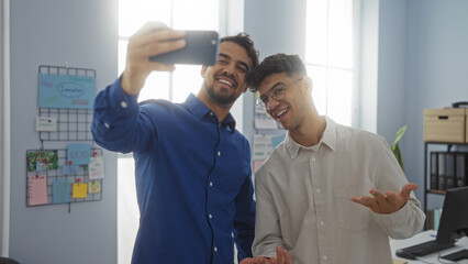 Fototapeta premium Hispanic men taking a selfie in an office, showcasing their work environment through a friendly and collaborative atmosphere.
