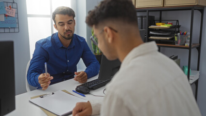 Fototapeta premium Men discussing project in office, one wearing blue shirt, another in white, surrounded by office supplies and documents, emphasizing teamwork in a business environment